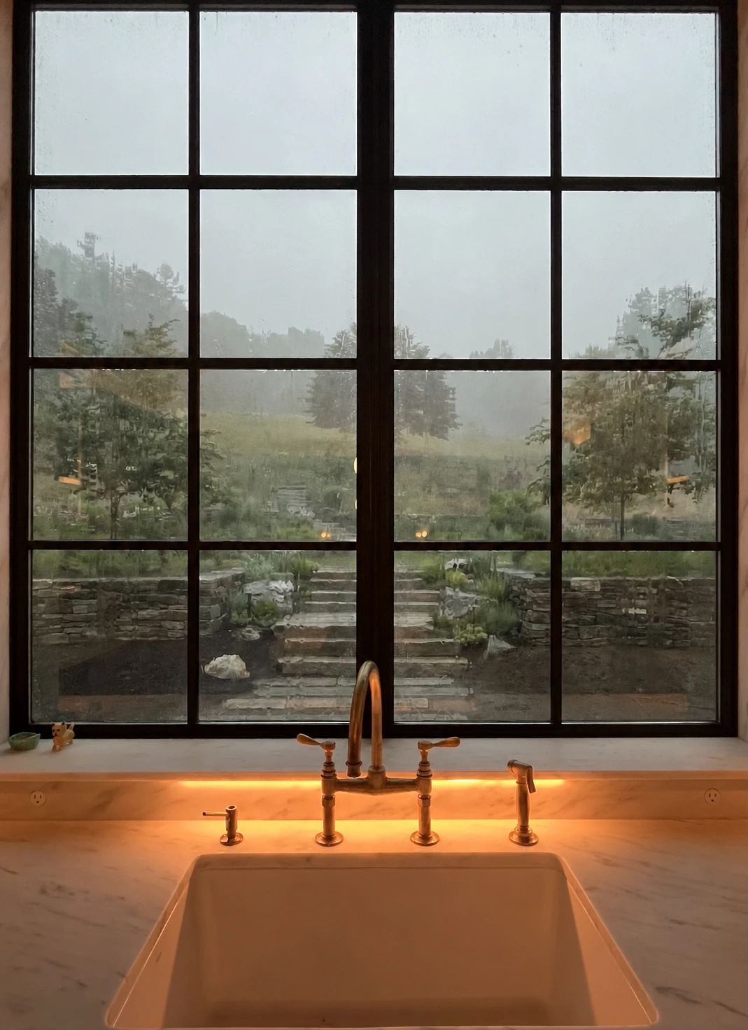 View from a kitchen window across stone garden walls and central steps toward a planted hillside in the rain